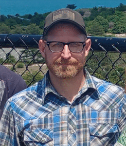 A photo of James, wearing a plaid shirt in tones of blue and grey and a baseball style cap.