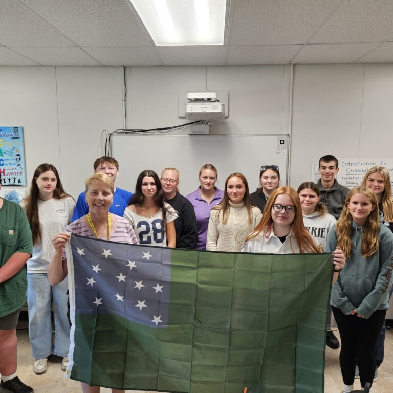 A number of students stand around an instructor and a smiling student holding a Green Mountain Boys flag