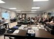Students sit around dark brown tables looking at a display board with information on it. The board can not be seen clearly in the photo.