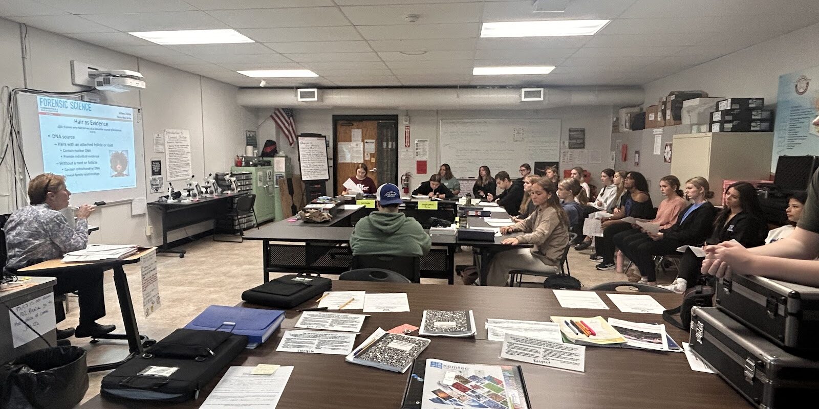 Students sit around dark brown tables looking at a display board with information on it. The board can not be seen clearly in the photo.