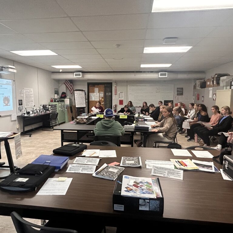 Students sit around dark brown tables looking at a display board with information on it. The board can not be seen clearly in the photo.