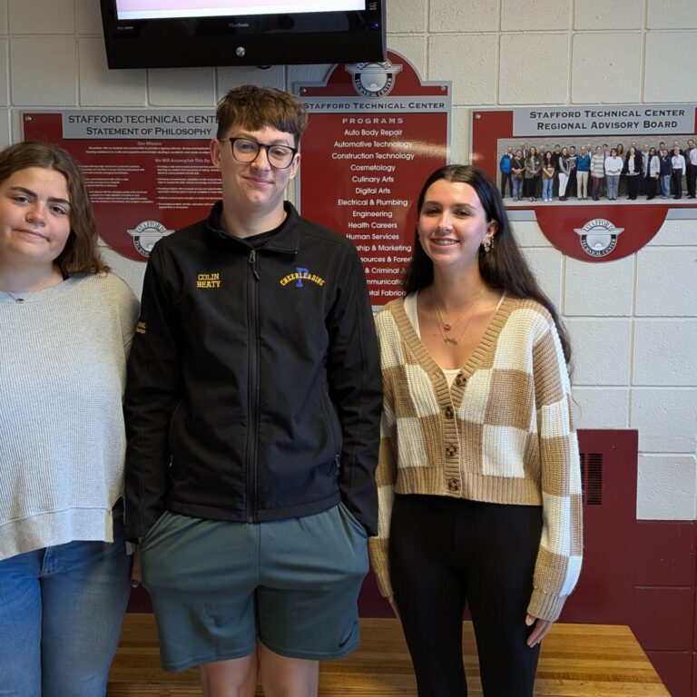 Piper, Colin, and Taylor smile at the camera. They are standing in front of a cinderblock wall