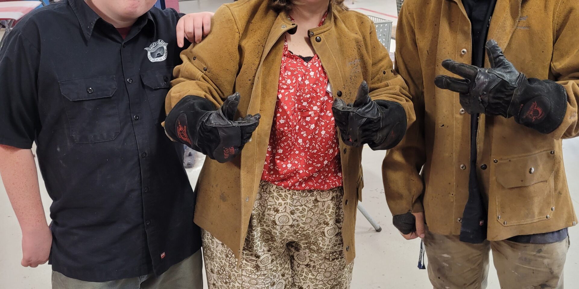 Two students stand on either side of school counselor. Counselor and one student are wearing welding jackets, and welding helmets. The helmets are flipped up so we can see the faces. In the background a red car is up in the air on an automotive lift.