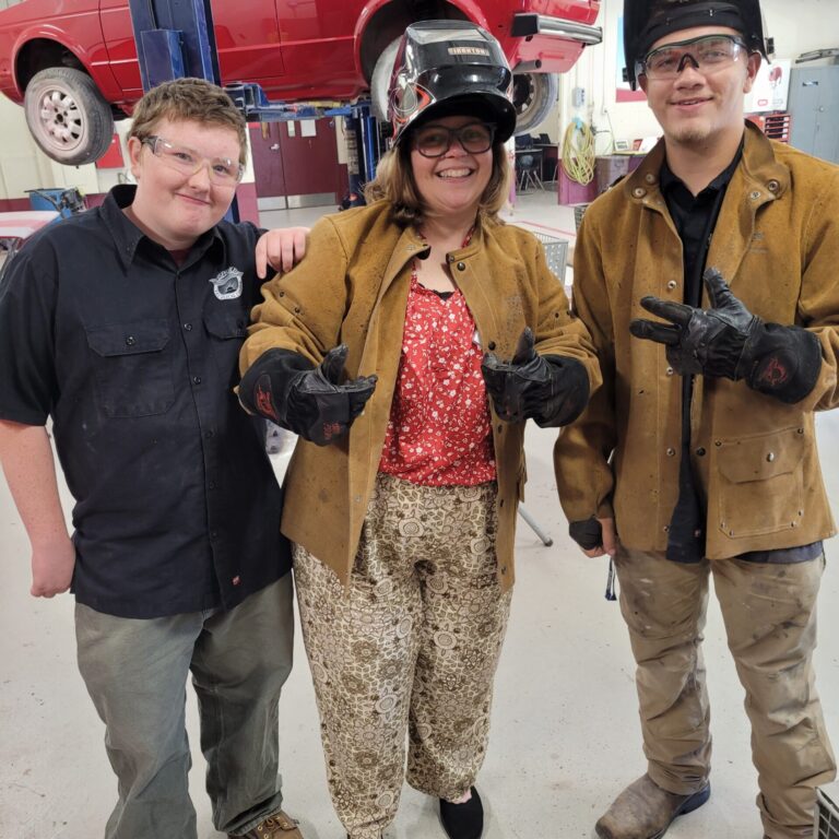 Two students stand on either side of school counselor. Counselor and one student are wearing welding jackets, and welding helmets. The helmets are flipped up so we can see the faces. In the background a red car is up in the air on an automotive lift.
