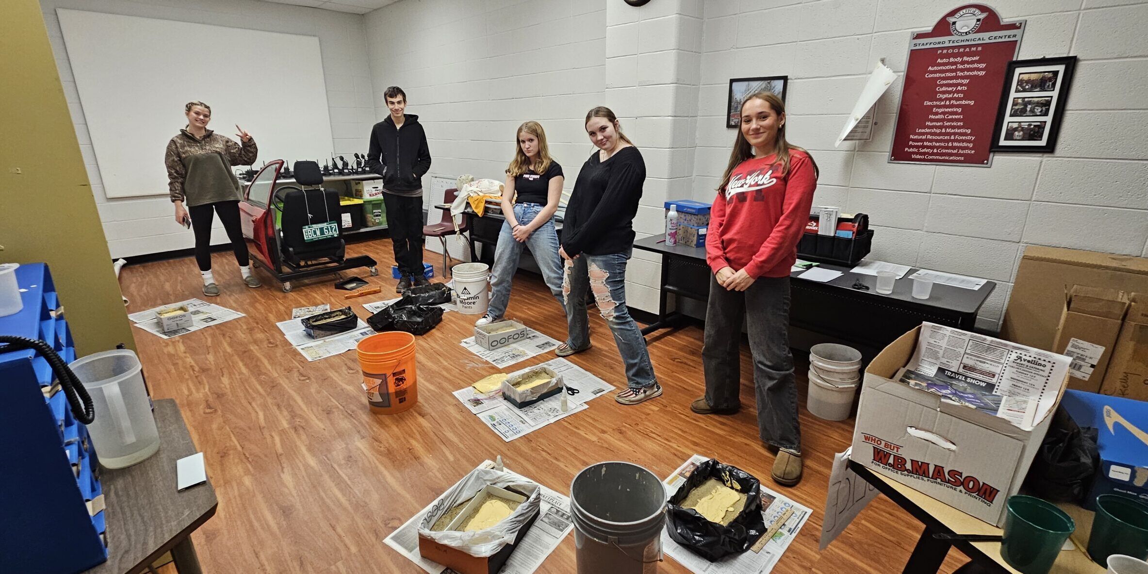 20251024_110849 – J Kelly, STC PS-CJ 5 Students stand around the perimeter of a room. On the floor in front of each of them is spread out newspaper. On the newspaper is a cast of their shoe print.