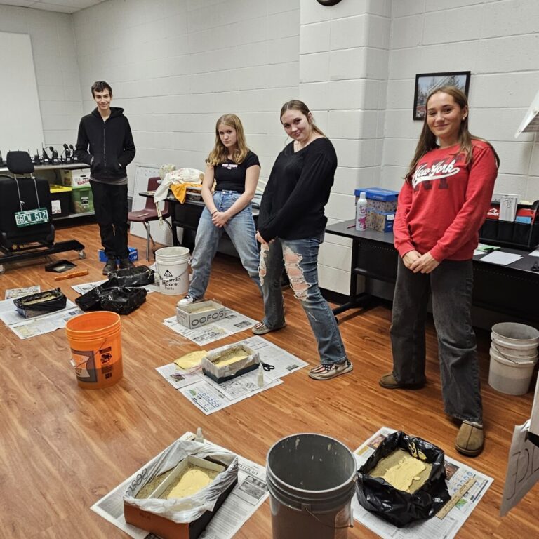 20251024_110849 – J Kelly, STC PS-CJ 5 Students stand around the perimeter of a room. On the floor in front of each of them is spread out newspaper. On the newspaper is a cast of their shoe print.