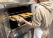 A student is pulling a tray of baked items out of an oven. We can only see the side of his body and face