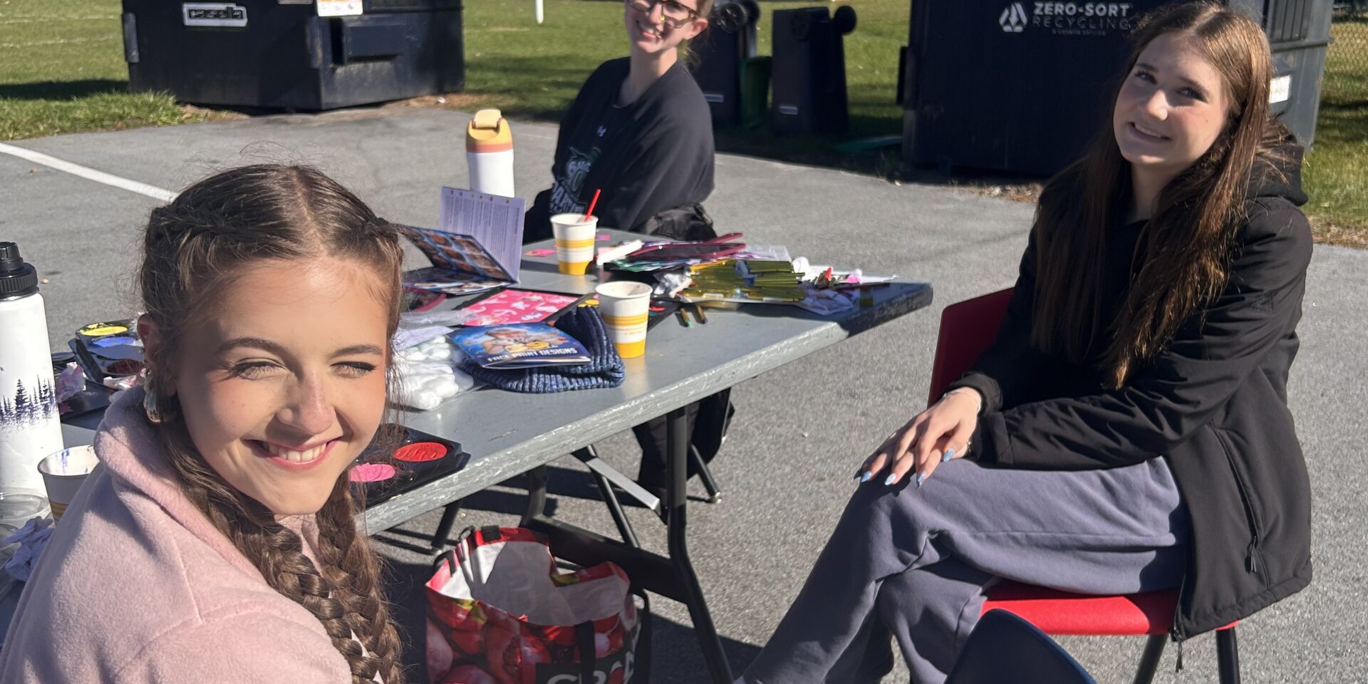 Three students sit around a table. All are smiling at the camera, the table has face painting materials.