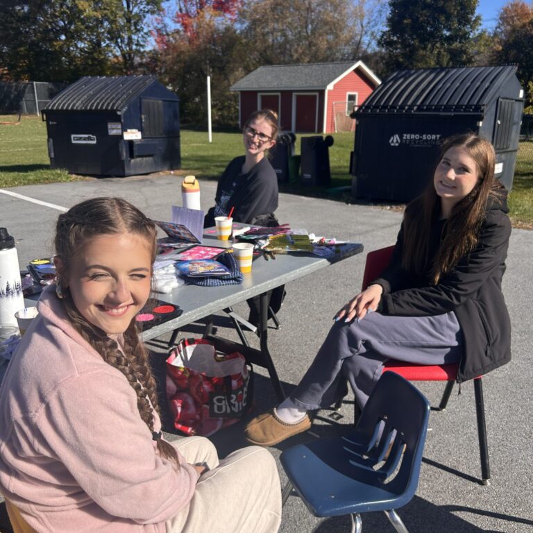 Three students sit around a table. All are smiling at the camera, the table has face painting materials.