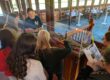 7 students surround a diorama of a ship in a glass case. Ons student has clipboard, and two others are holding measuring tapes.