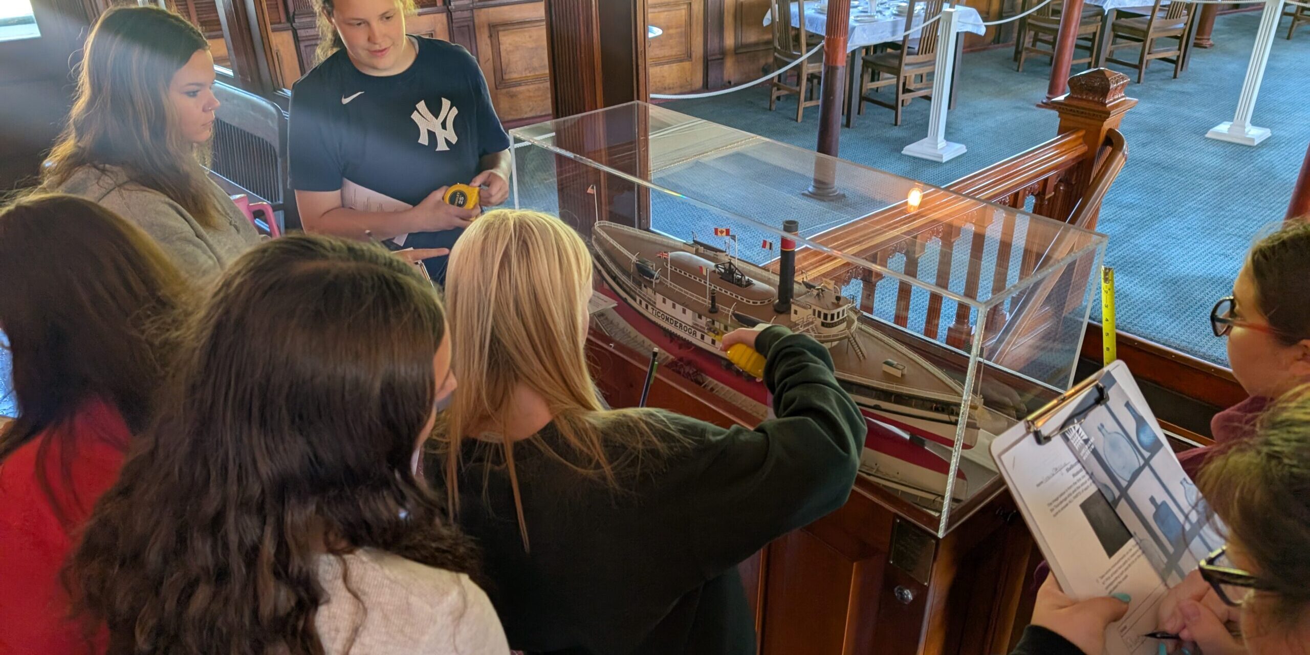 ticonderoga trip 2025 7 students surround a diorama of a ship in a glass case. Ons student has clipboard, and two others are holding measuring tapes.