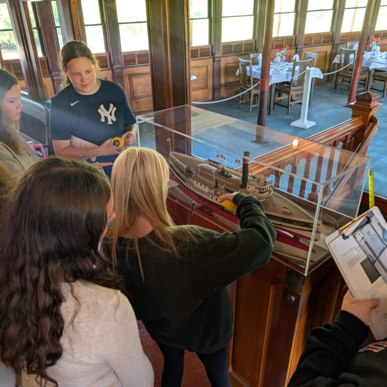 7 students surround a diorama of a ship in a glass case. Ons student has clipboard, and two others are holding measuring tapes.