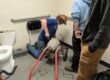 One student is operating a sewer cleaning machine while two other students look on. The toilet has been removed from its location and is off to the side.