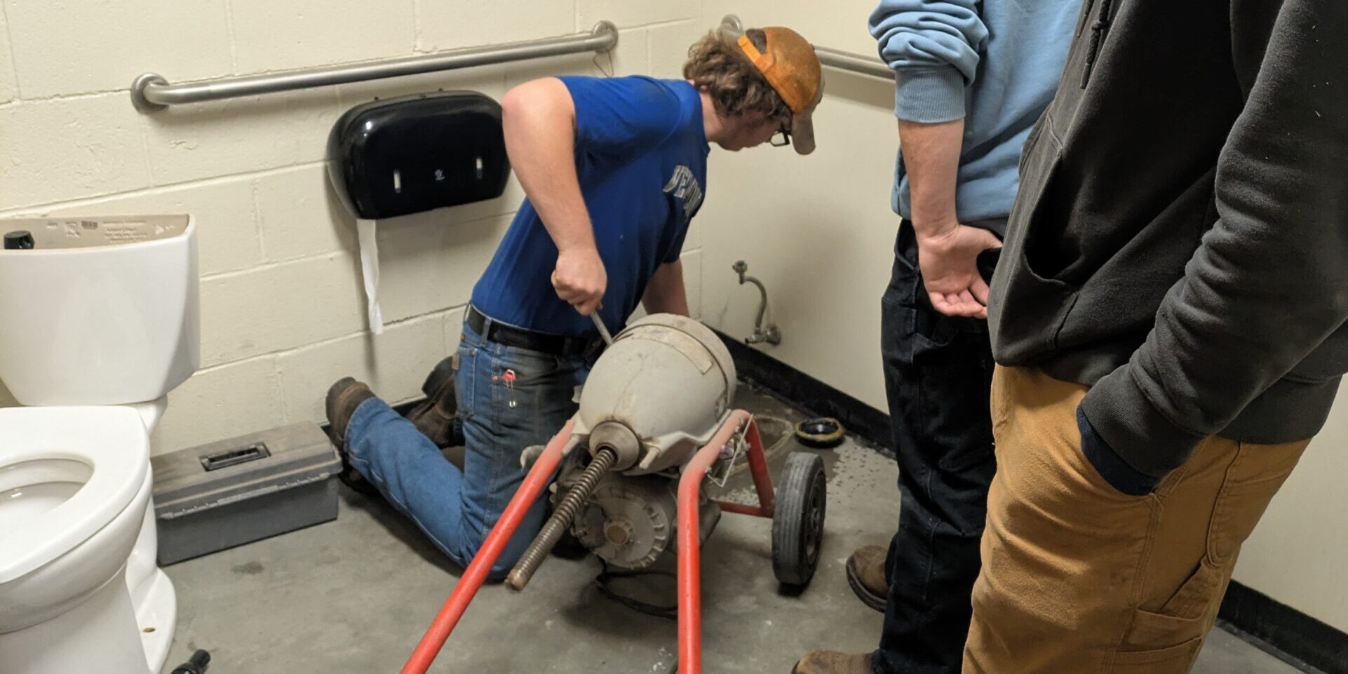 One student is operating a sewer cleaning machine while two other students look on. The toilet has been removed from its location and is off to the side.