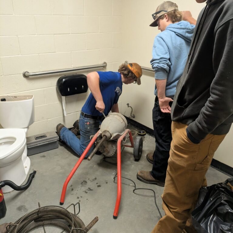 One student is operating a sewer cleaning machine while two other students look on. The toilet has been removed from its location and is off to the side.