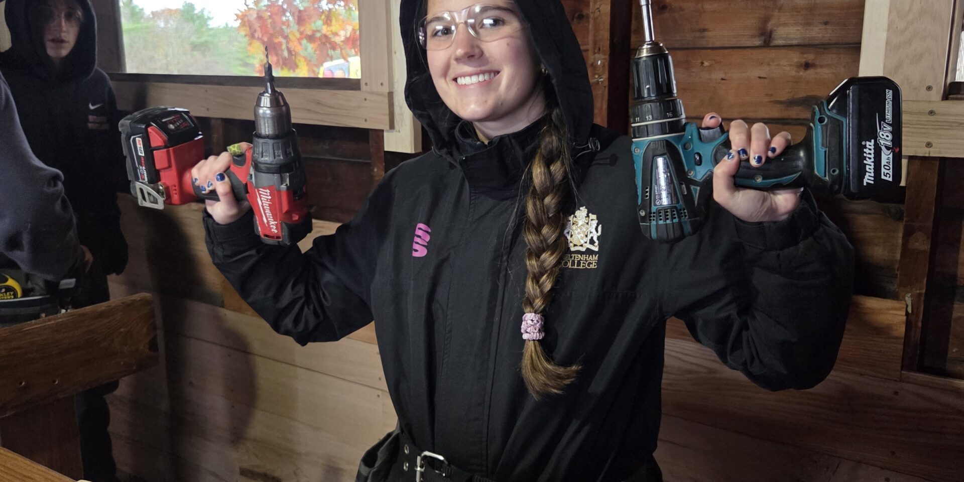 20251030_122143 – Tyler Gugliotta A young lady holds two cordless drill drivers, one in each hand. She is standing in front of some cut boards that are on a table.