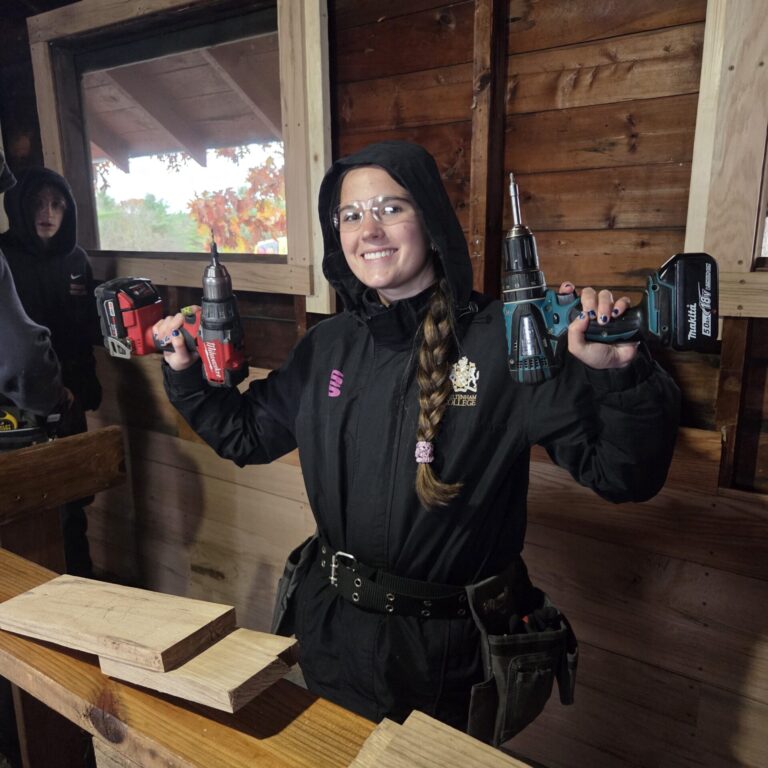 20251030_122143 – Tyler Gugliotta A young lady holds two cordless drill drivers, one in each hand. She is standing in front of some cut boards that are on a table.