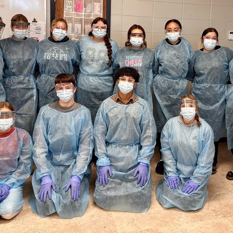 12 Students dressed in blue gowns, wearing purple gloves, and face masks face the camera