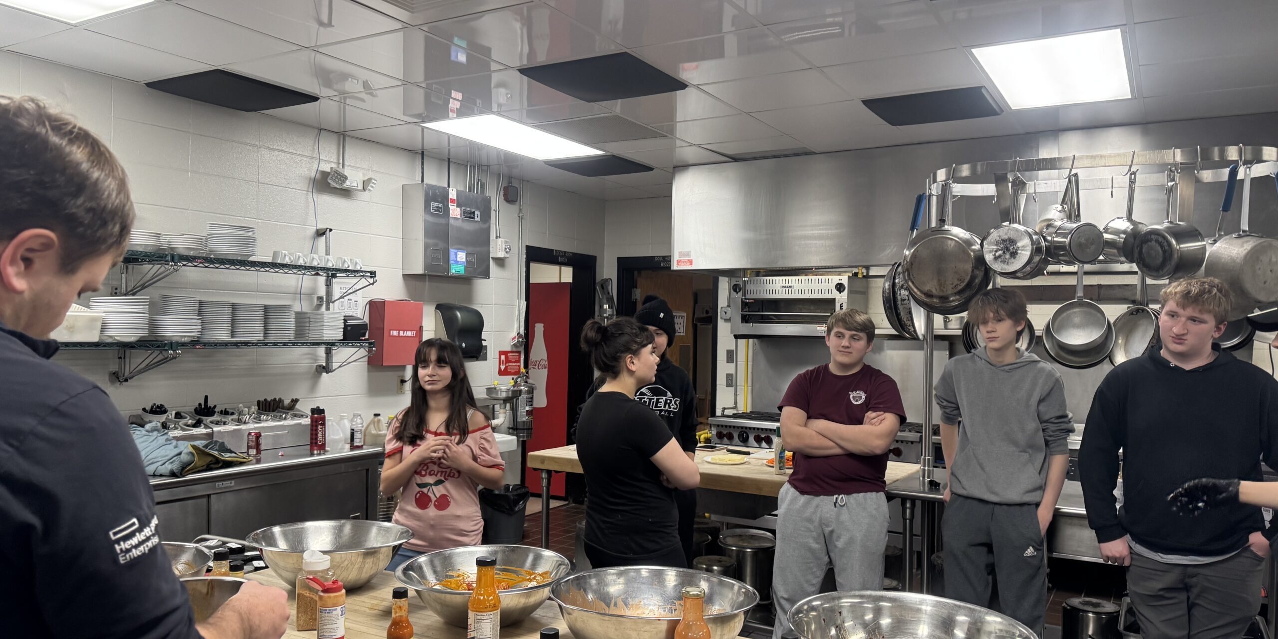 culinary studetns stand in front of a work station with multiple metal bowls filled with hot sauce, waiting to toss fried chicken wings into the bowls