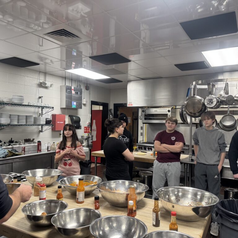 culinary studetns stand in front of a work station with multiple metal bowls filled with hot sauce, waiting to toss fried chicken wings into the bowls