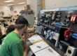 two students sit in front of an upright board covered with control switches.