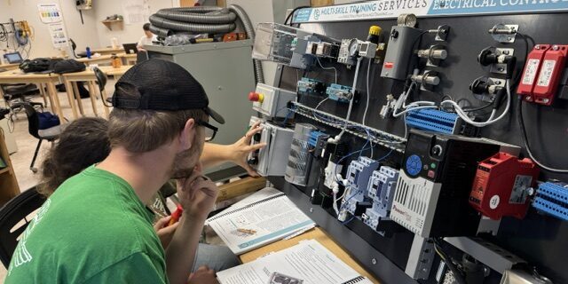 two students sit in front of an upright board covered with control switches.