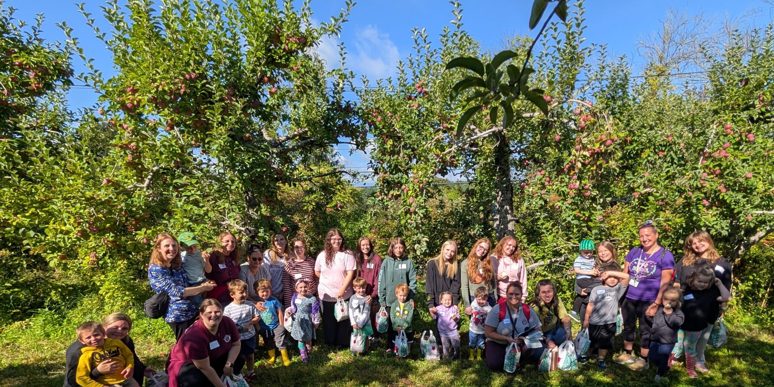 PXL_20260918_143908457~2 – Jacqueline Hemple A group of adults, high school students and preschoolers stand in front of fruit laden apple trees.