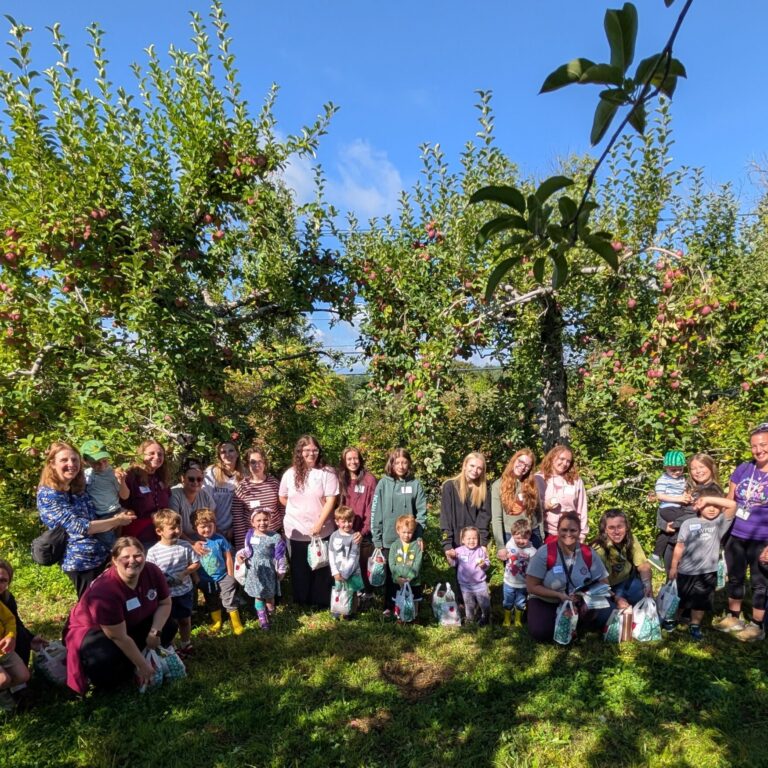 PXL_20260918_143908457~2 – Jacqueline Hemple A group of adults, high school students and preschoolers stand in front of fruit laden apple trees.