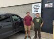 Two men stand next to a vehicle, behind them on the wall of a building is a sign that read, Vermont Official Inspection station
