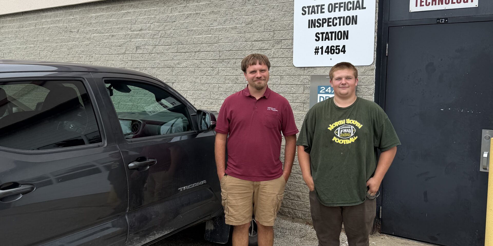 IMG_0313 (1) – Eslie Jones Two men stand next to a vehicle, behind them on the wall of a building is a sign that read, Vermont Official Inspection station