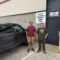 Two men stand next to a vehicle, behind them on the wall of a building is a sign that read, Vermont Official Inspection station