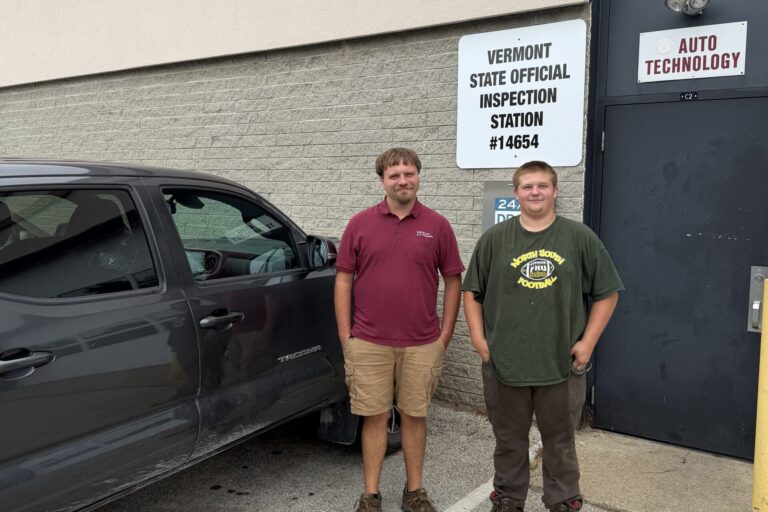 Two men stand next to a vehicle, behind them on the wall of a building is a sign that read, Vermont Official Inspection station