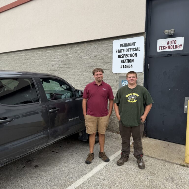 IMG_0313 (1) – Eslie Jones Two men stand next to a vehicle, behind them on the wall of a building is a sign that read, Vermont Official Inspection station